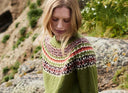 A woman wearing a green knit sweater with a multicolor Fair Isle yoke pattern and a dark navy skirt, posing outdoors on a rocky, grassy hillside.